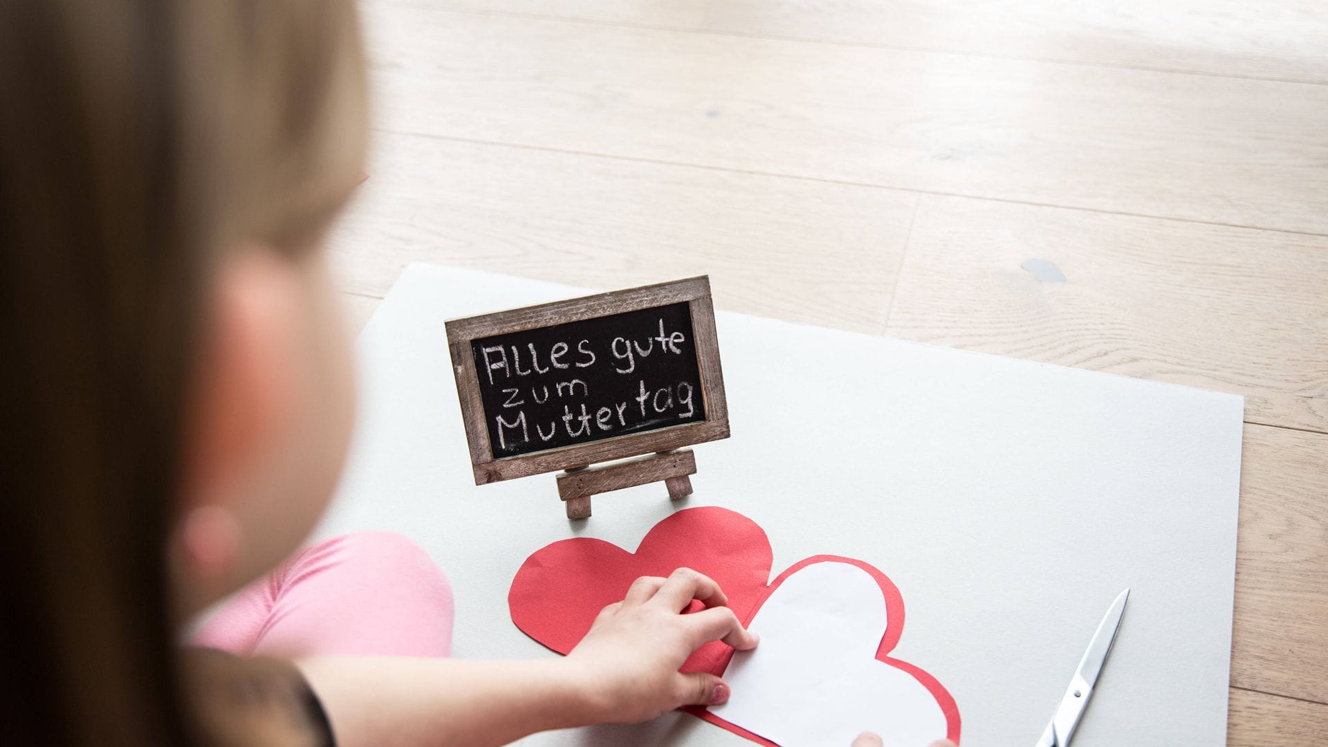 Ein kleines Mädchen sitzt auf einem Laminatfussboden und bastelt zum Muttertag für die Mutter ein Herz aus Pappe als Geschenk, daneben liegt eine Schere. Auf einer Tafel steht mit Kreide geschrieben, alles gute zum Muttertag.
