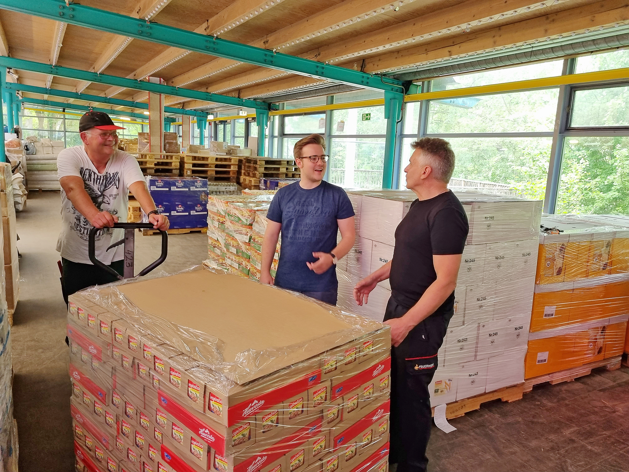 Помічник-волонтер Хольгер Старк (зліва) везе новий піддон з продуктами на склад у Клостерзеехалле, де Максиміліан Рейнхардт і пожежний Пітер Нойманн оглядають інвентар. Обидва погоджуються, що «Допомога замість хом’яків» терміново потребує додаткових пожертвувань від населення, щоб мати можливість забезпечити людей України та біженців з війни, які приїжджають сюди, гуманітарною допомогою у перевіреному вигляді.   Зображення: Гаманн.